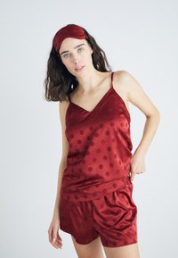 Pijamale din satin roșu, cu un top tip tank cu decolteu în V și puncte polka, însoțite de șorturi asortate. Acestea vin împreună cu o mască de somn din satin coordonată.