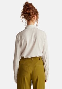 Camicia a righe in tessuto leggero, con colletto e polsini abbottonati, abbinata a pantaloni sartoriali verde oliva, con dettagli sulle tasche posteriori.