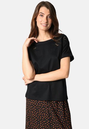 Femme aux longs cheveux bruns portant un haut noir à manches courtes avec des découpes florales et une jupe noire avec des taches orange, souriant doucement.