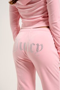 Rozā velūra treniņtērps ar akmens detaļām uz muguras, kas rotāts ar vārdu "Juicy". Materiāls ir mīksts un gluds.