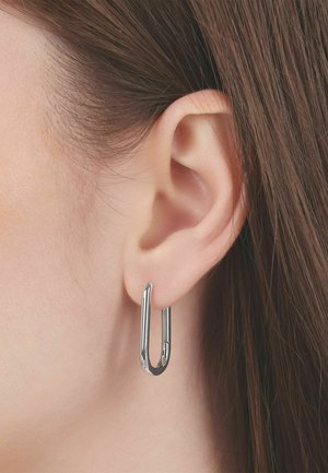 Primer plano de una oreja con un pendiente de aro alargado ovalado, de tono plateado y recto, llevado en el lóbulo, con un fondo de cabello castaño.