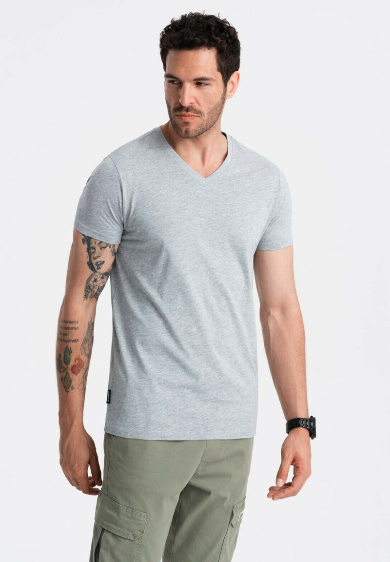 Šedé tričko s v-neck výstřihem vyrobené z měkké látky, krátké rukávy a běžný střih, v kombinaci s olivově zelenými cargo kalhotami. Viditelné tetování na pažích.