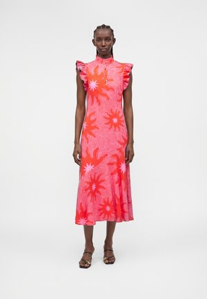 Frau steht und trägt ein pink-rot ärmelloses Midikleid mit Rüschenschultern und Sonnenstrahlenmuster, kombiniert mit schwarzen offenen Sandalen.