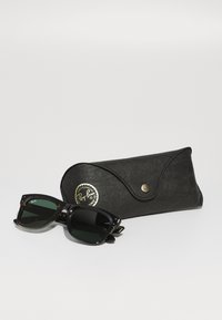 Ochelari de soare Ray-Ban cu lentile de culoare verde închis, ramă de tip țestoasă și accente metalice, însoțiți de un etui negru texturat cu un logo aurit.