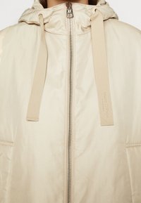 Veste à capuche beige avec fermeture éclair argentée à l'avant et larges cordons, un cordon avec le nom de la marque embossé verticalement.