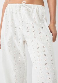 Vita broderade byxor i lätt tyg som har ögonblomsmönster, en elastisk midja och dekorativ dragsko.