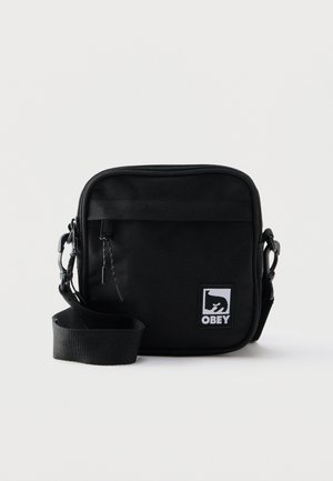 Zwarte schoudertas in vierkante vorm met ritsvak aan de voorkant, verstelbare riem en witte Obey-logo patch rechtsonder op de voorkant.
