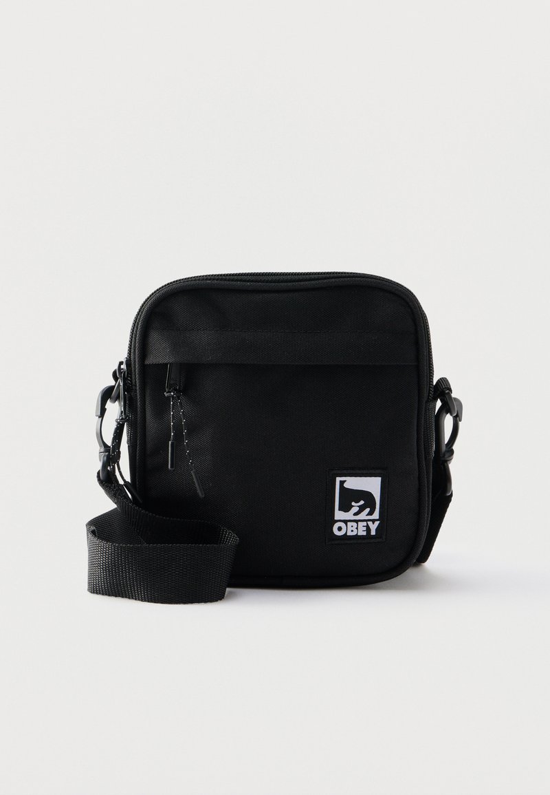 Czarna torba na ramię w kształcie kwadratu, z przednią kieszenią na zamek, regulowanym paskiem oraz białą naszywką z logo Obey na przednim, dolnym prawym rogu.