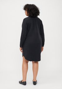 Black shirt dress with long sleeves, straight cut, side slits, and a buttoned cuff. Soft fabric with a smooth texture. Worn with studded black flats.