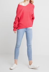 Kirkas vaaleanpunainen ylipitkä neule, jossa on rento muotoilu, yhdistettynä vaaleansinisiin skinny-farkkuihin ja valkoisiin tennareihin. Rakenteellinen neulekangas.