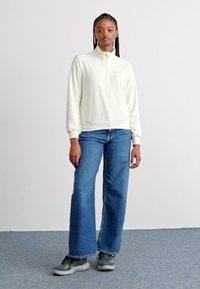 White, zip-neck sweatshirt with a relaxed fit, paired with light blue, wide-leg jeans and gray and green sneakers. Simple, textured fabric.