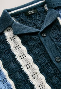 Stickad pikétröja i marinblått med vita och grå texturerade ränder. Har en krage och två knappar framtill. Tyget är andningsbart.