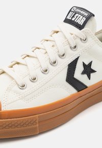 Baskets montantes en toile de couleur crème, présentant un logo étoile noir, une semelle en caoutchouc orange et des œillets métalliques avec des lacets blancs.