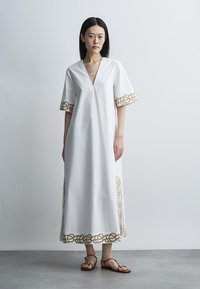 Femme debout portant une robe maxi blanche avec broderie chaîne dorée sur les manches, l'ourlet et les côtés, associée à des sandales plates marron.