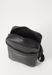 Černá kožená crossbody taška se zipem nahoře, přední kapsou a logem. Vlastnosti zahrnují texturovaný povrch a nastavitelné ramenní popruhy.