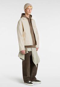 Beige Baumwolljacke über einem braunen Hoodie, kombiniert mit braunen weiten Hosen und schwarz-weißen gemusterten Schuhen. Hellgrünes Hemd um die Taille gebunden.