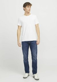 Weißes Baumwoll-T-Shirt, Rundhalsausschnitt, kombiniert mit dunkelblauen Jeans. Modell steht. Weiße Sneaker mit schwarzen Akzenten an der Seite.