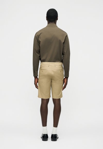 Homme de dos portant une chemise kaki à manches longues, un short beige aux genoux, des chaussettes blanches et des chaussures de ville noires sur un fond uni.
