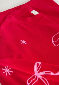 Červené bavlnené tričko poskladané s okrúhlym výstrihom, s ružovými grafickými potlačami hviezd a detailom motýlika. Hladká textúra a živá farba.