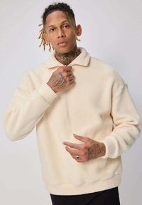 Pull en polaire couleur crème, avec un col, des manches longues et une texture douce, offrant une coupe décontractée.