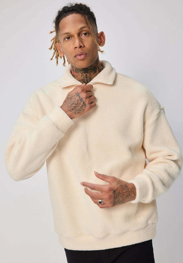Pull en polaire couleur crème, avec un col, des manches longues et une texture douce, offrant une coupe décontractée.