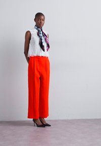 Hvid blonder sleeveless top, lysorange brede bukser og et mønstret tørklæde. Model iført sorte højhælede sko, stående foran en neutral baggrund.