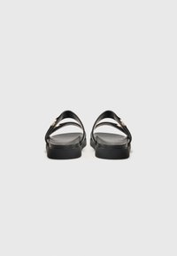 Black sandals with two curved straps and a flat sole, featuring a subtle texture and a small gold accent near the straps.