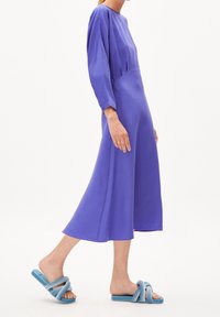 Lungo vestito viola con scollatura rotonda e maniche lunghe, abbinato a sandali blu alla moda con cuciture a spirale e suole testurizzate.