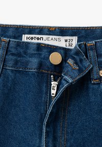 Jeans de mezclilla en azul oscuro con costuras naranjas, con un botón de metal, cierre de cremallera y etiquetas que indican el tamaño W27 L32.