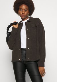 Σκούρο καφέ, υφασμάτινο, πουκάμισο με κουμπιά και δύο μπροστινές τσέπες. Φορεμένο πάνω από λευκό μπλουζάκι, σε συνδυασμό με μαύρα δερμάτινα παντελόνια και μια μαύρη τσάντα.