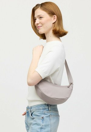 Jonge vrouw met kort rood haar poseert zijwaarts, draagt een witte top met korte mouwen, lichtblauwe jeans en draagt een lichtgrijze schoudertas in halvemaanvorm.