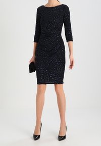 Un vestido azul marino con mangas tres cuartos, silueta ajustada, patrón texturizado y escote redondo. Combinado con un bolso de mano negro y tacones.