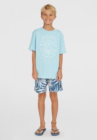 Šviesiai mėlynas medvilninis marškinėlis su baltu grafiniu spaudiniu. Tamsiai mėlynos šortai su baltu lapų raštu. Mėlynos šlepetės. Kasdienė paplūdimio apranga.