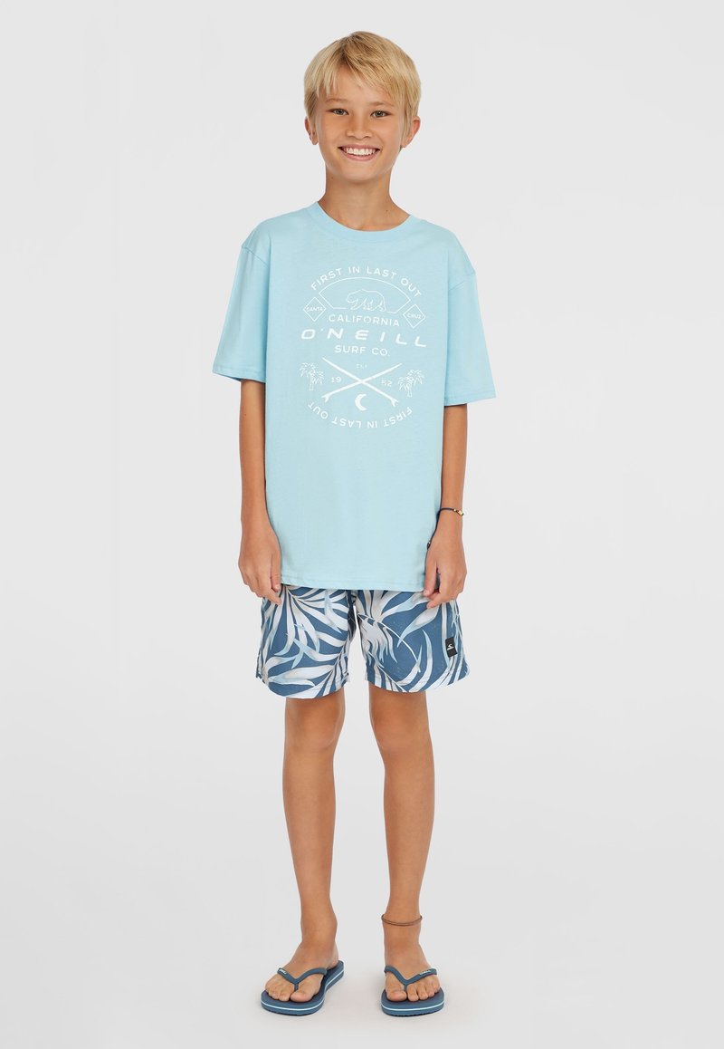 Šviesiai mėlynas medvilninis marškinėlis su baltu grafiniu spaudiniu. Tamsiai mėlynos šortai su baltu lapų raštu. Mėlynos šlepetės. Kasdienė paplūdimio apranga.