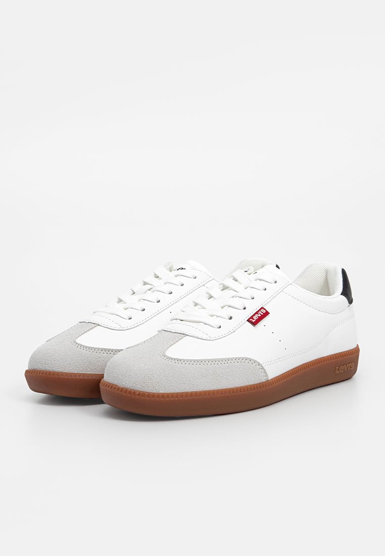 Zapatillas blancas con acentos de ante gris, parte superior de cuero y suela de goma marrón. Presentan un sistema de cordones y una etiqueta de logo roja en el costado.