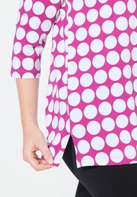 Top fucsia con grandi pois bianchi, maniche a tre quarti, fessure laterali e vestibilità morbida; mostrato mentre viene tirato all'orlo.
