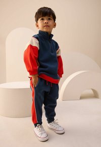 Blå og rød fleece træningsdragt med lynlås ved halsen, kontrasterende ærmer, elastiske manchetter og sidelister; parret med hvide sneakers.