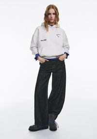 Witte hoodie met een voorzak en geborduurde details, gecombineerd met brede zwarte spijkerbroeken en grijze laarzen met blauwe veters.