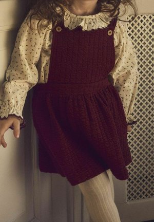 Vestido rojo con textura y falda fruncida, escote redondo adornado con botones, sobre una blusa crema con estampado floral, combinado con medias blancas.