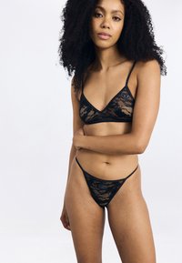 Ensemble de lingerie en dentelle noire comprenant un bralette à fines bretelles et un string assorti. Motif floral avec des détails matériels minimalistes.