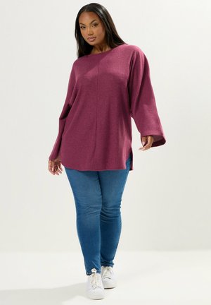Maglione oversize fucsia lavorato a maglia con maniche lunghe, spalle basse, spacchi laterali, indossato con jeans blu aderenti e sneakers bianche.