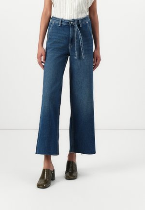 Femme portant un jean bleu taille haute à jambes larges avec ourlets effilochés, ceinture nouée, blouse blanche sans manches et mules à talons foncées.