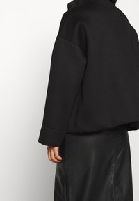 Černá oversized mikina s rovnými lemy, spadlými rameny a širokými manžetami. Měkká textura, nošena přes černou koženou sukni.