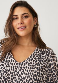 Een blouse met luipaardprint en V-hals, met korte pofmouwen. Het materiaal heeft een geribbelde textuur in tinten van beige en zwart. Zilveren oorbellen.