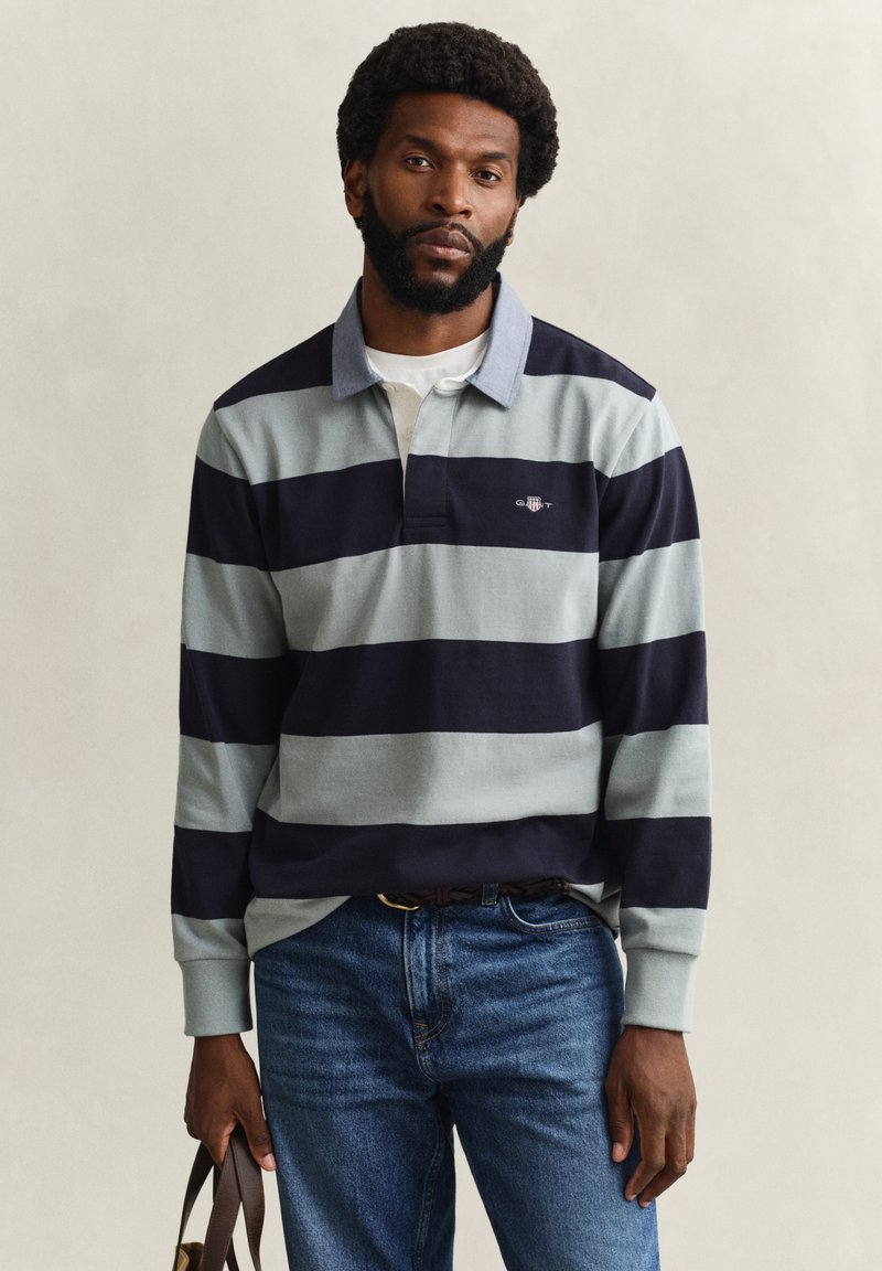 Uomo con afro e barba che indossa una maglia da rugby a righe blu navy e grigio chiaro e jeans, che tiene in mano sinistra una cintura di pelle marrone.