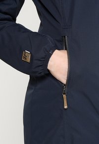 Mano en el bolsillo con cremallera de una chaqueta azul marino con puño elástico y un pequeño parche de logo marrón en la manga.