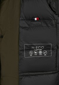 Intérieur d'une veste matelassée verte avec doublure noire, une poche révélant un accent tricolore et une étiquette détaillant des matériaux durables.