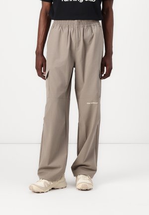 Personne portant un pantalon cargo beige avec une taille élastique et le logo « new balance » sur la jambe gauche, assorti à des chaussures de sport blanches.