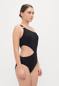 Maillot de bain en une pièce noir avec un design découpé, une bretelle, et un accent en matériel circulaire. Texture lisse et coupe ajustée.
