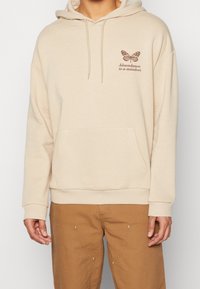 Sudadera con capucha beige con bolsillo frontal, capucha con cordón y un gráfico de mariposa marrón con texto en el pecho. Material de mezcla de algodón.